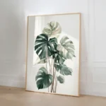 Large framed art print of a Monstera plant, leaning on the floor against a white wall with wainscoting details in a bright, elegant living room.