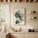 Framed art print showcasing a lush Monstera plant, adorning a light wall above wooden shelves and countertops in a modern, neutral-toned kitchen.