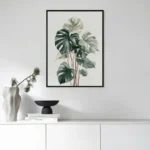 Black framed art print of a Monstera plant, hung on a white wall above a modern white console table with a vase and decorative items in a living room.