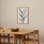 Framed black and white tropical leaf line art print, adorning a light-colored wall in a bright and airy dining area with simple wooden furniture.