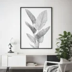 Black framed print of black and white tropical leaf line art, hung above a white sideboard with a green plant in a contemporary living room.