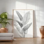Large framed print of black and white tropical leaf line art, leaning on the floor against a white wall, accompanied by a potted green plant.