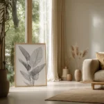Large framed print of black and white tropical leaf line art, leaning against a light wall by a tall window with lush green plants in a cozy living room.