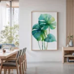 Vibrant watercolor art print of blue and green monstera leaves, framed and hung on a white wall in a bright, modern dining room with a wooden table and chairs.