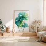 Large framed watercolor art print of tropical monstera leaves, leaning on the floor against a light wall in a minimalist bedroom with natural light and dried grass decor.