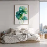 Framed watercolor art print of monstera leaves in shades of green and blue, hung on a white wall above a bed in a sunlit modern bedroom with city views.