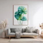 Framed botanical watercolor print of green and blue monstera leaves, displayed on a light wall above a simple grey sofa in a contemporary living room.