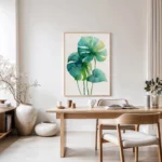 Framed watercolor art print featuring green and blue monstera leaves, displayed on a light wall above a wooden desk and chair in a minimalist home office or dining area.