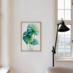 Framed watercolor art print of green and blue monstera leaves, hung on a white wall next to a large window and a black floor lamp in a minimalist room.